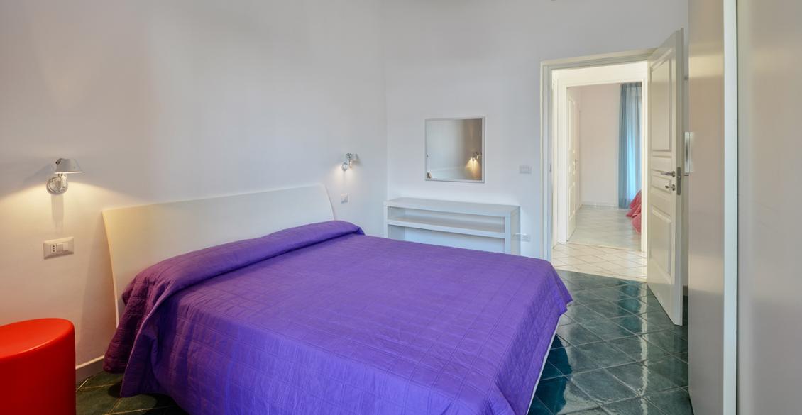 Camera luminosa, letto con copriletto viola, atmosfera serena e accogliente, pavimento lucido.