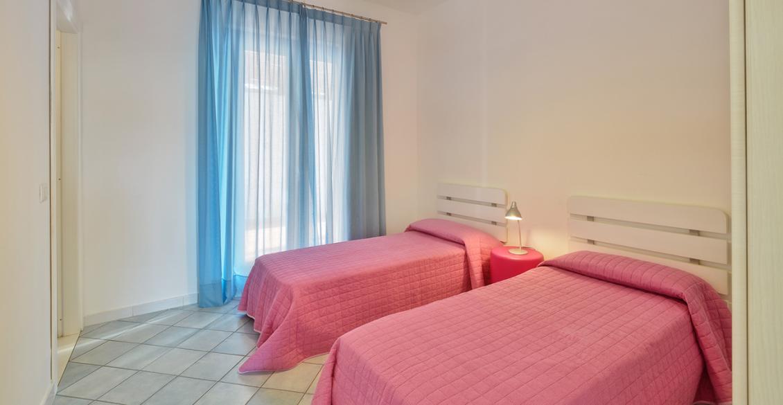 Camera luminosa con due letti singoli rosa e tende azzurre, atmosfera accogliente e serena.