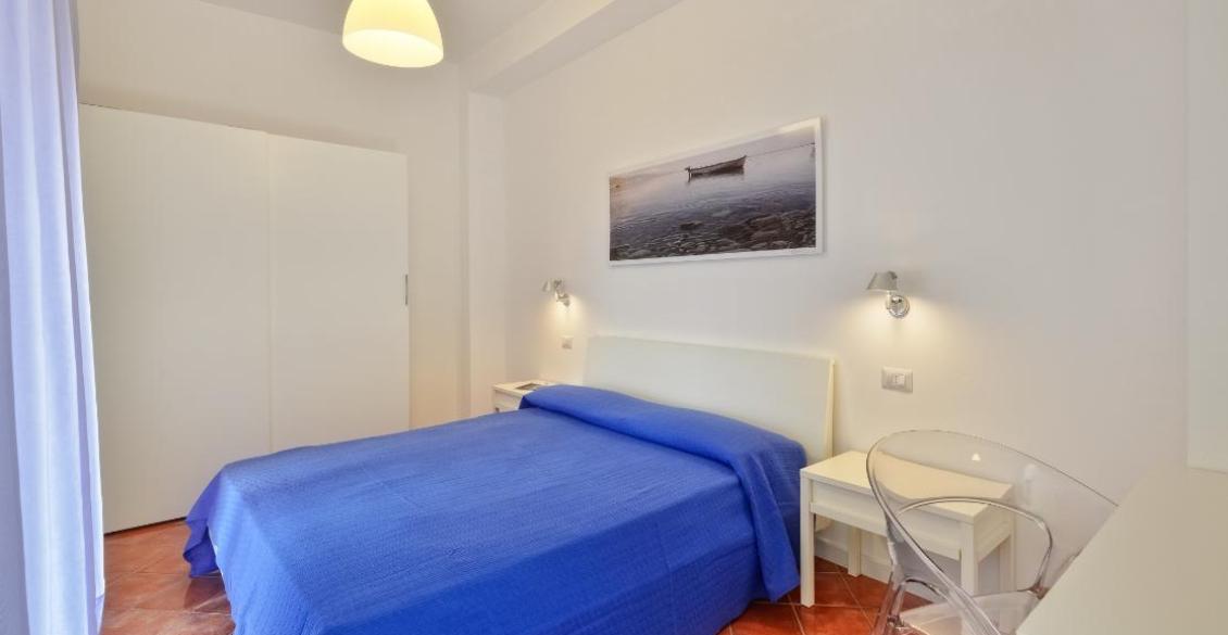 Camera luminosa, letto blu, atmosfera serena, sedia trasparente, quadro di barca sul mare.