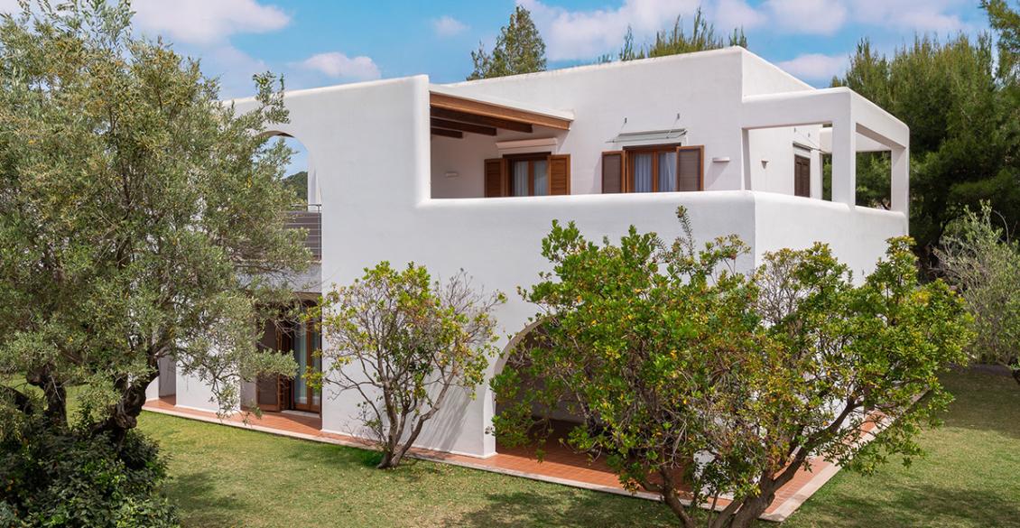 Villa moderna bianca con archi, immersa nel verde, sotto un cielo azzurro e sereno.