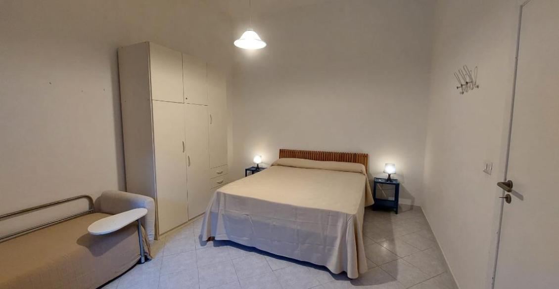 Camera semplice e accogliente, letto matrimoniale, armadio bianco, luce soffusa, atmosfera tranquilla.