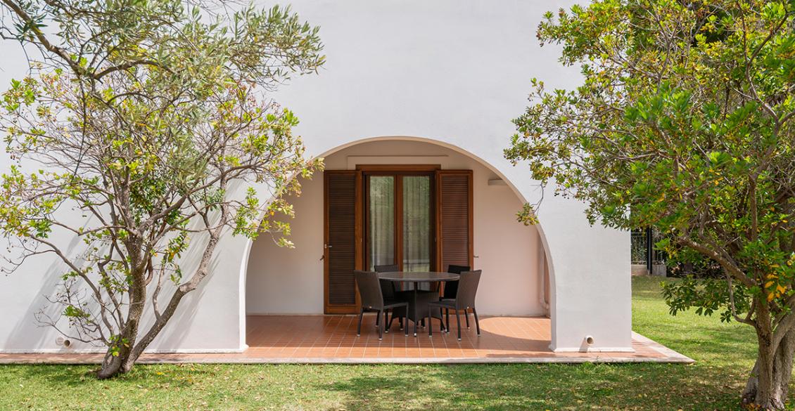 Veranda mediterranea con archi, sedie eleganti e natura rigogliosa.