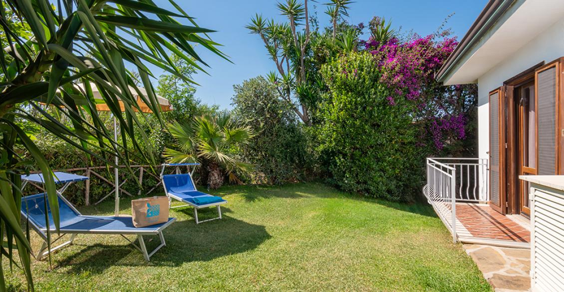 Giardino soleggiato con sdraio, ombrellone e casa accogliente, perfetto per rilassarsi all'aperto.