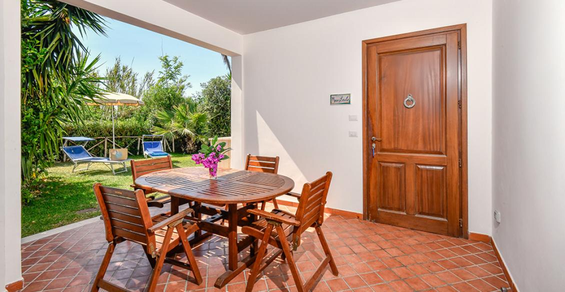 Veranda accogliente con tavolo in legno, circondata da verde rigoglioso e cielo sereno.
