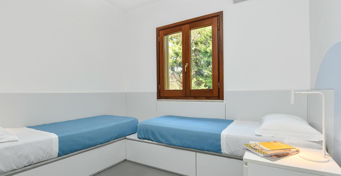 Camera minimalista con letti singoli, lenzuola azzurre, atmosfera serena e ordinata.