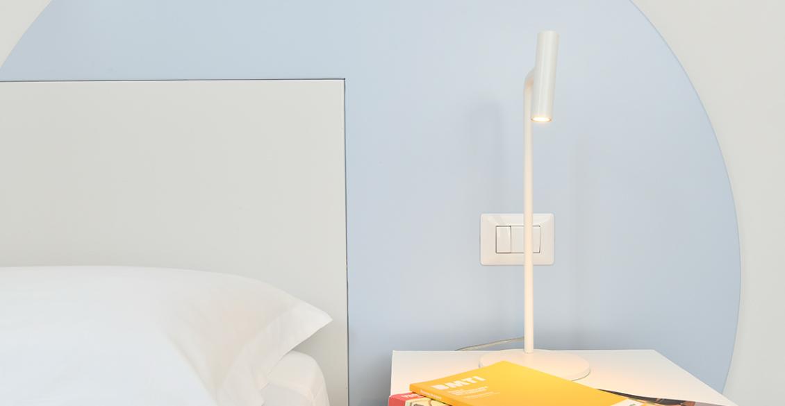 Camera minimalista: letto bianco, lampada moderna, libri colorati su comodino, atmosfera serena.