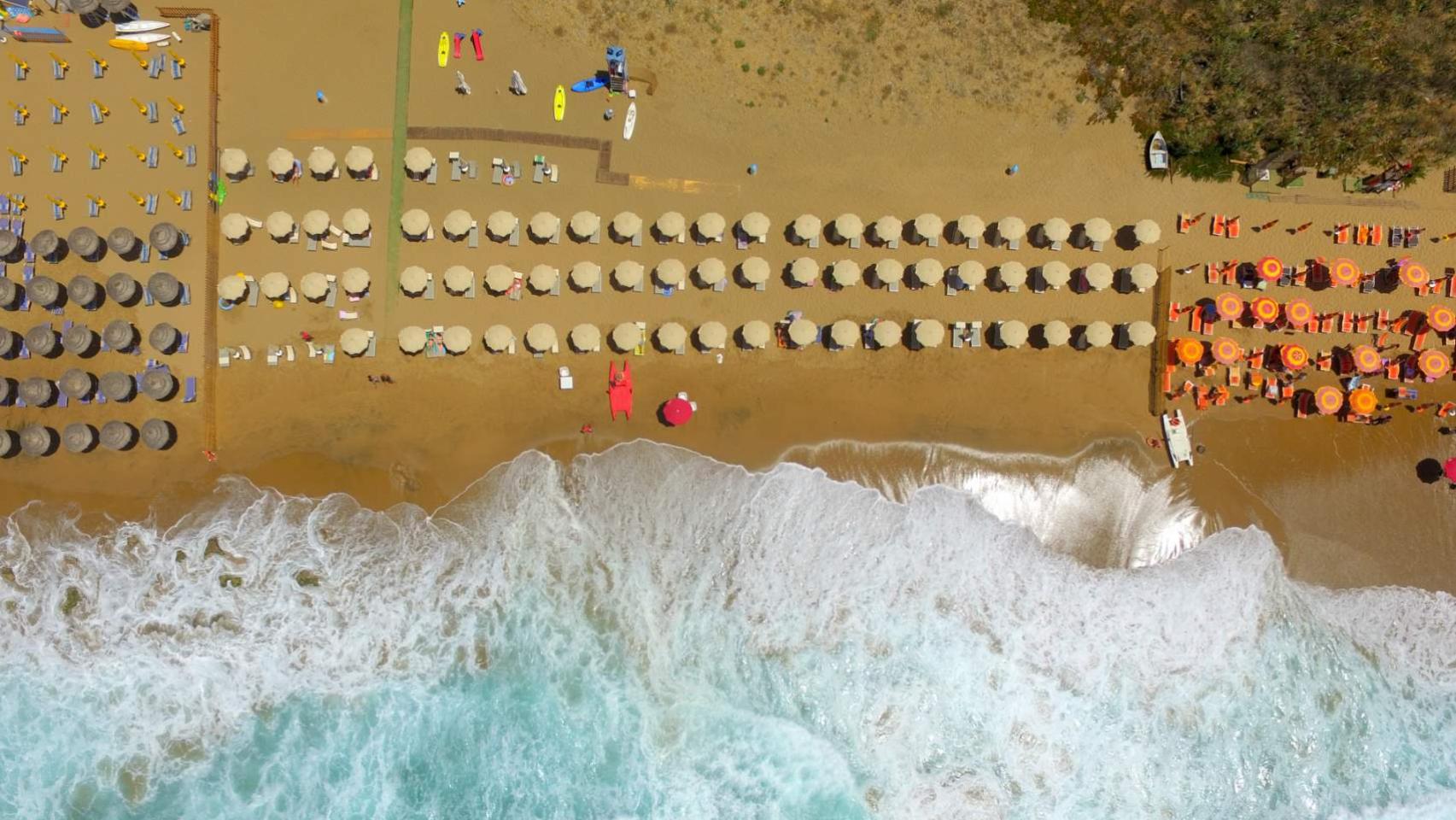 Vista aerea di una spiaggia con ombrelloni colorati e onde che si infrangono.
