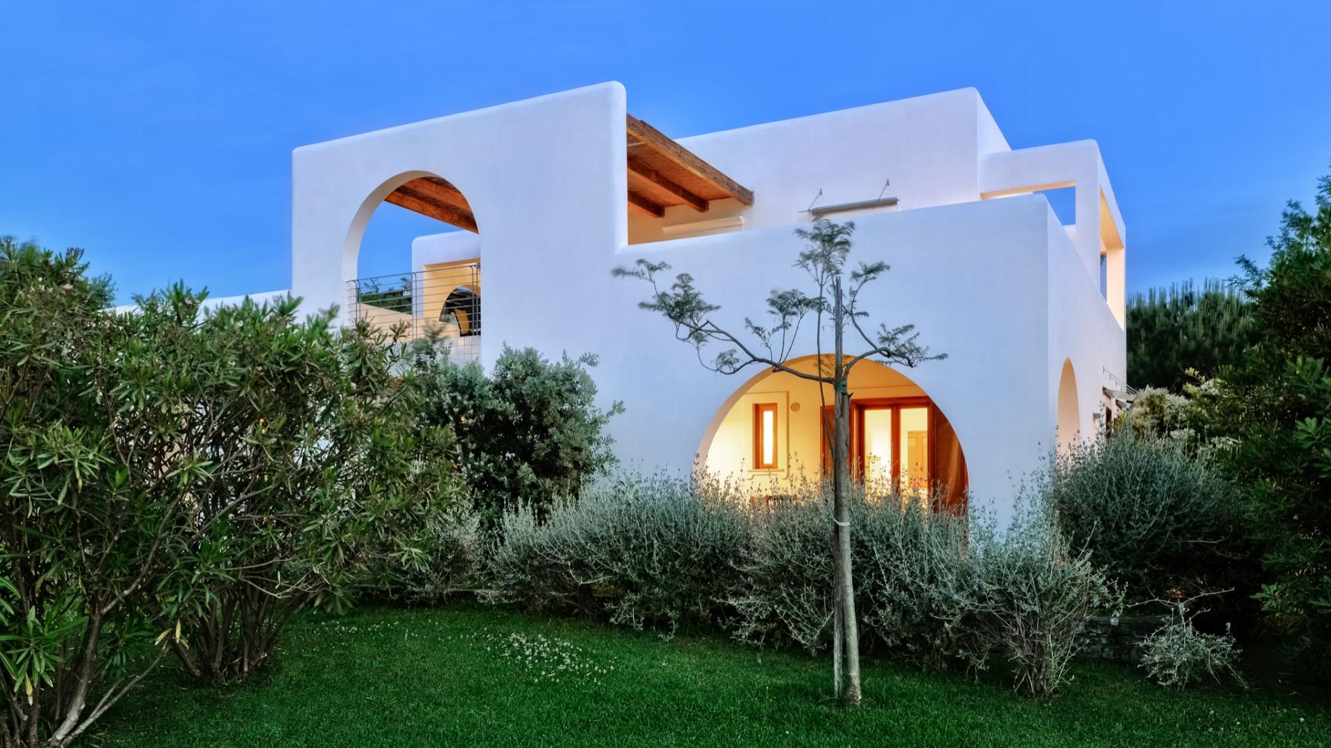 Casa bianca moderna con archi, circondata da verde rigoglioso, al tramonto sereno.