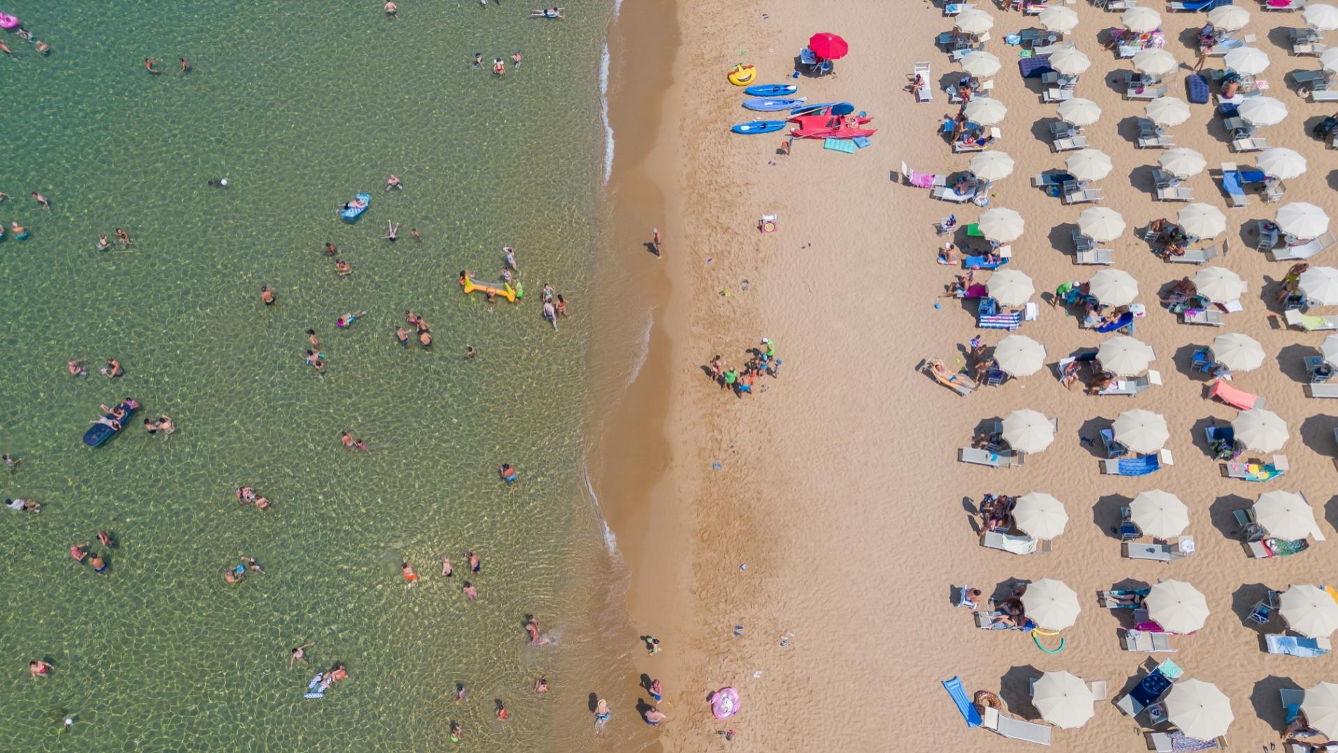 Spiaggia affollata, ombrelloni bianchi, mare cristallino, persone felici nuotano e si rilassano al sole.