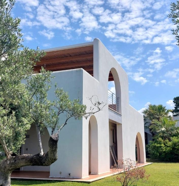 Villa moderna con archi bianchi, cielo azzurro e giardino verde rigoglioso, atmosfera mediterranea.