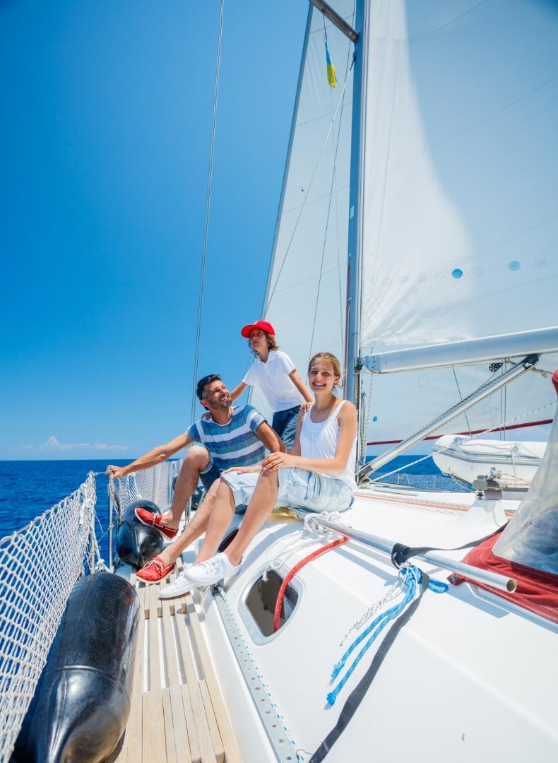 Famiglia sorridente su una barca a vela, cielo azzurro e mare scintillante.