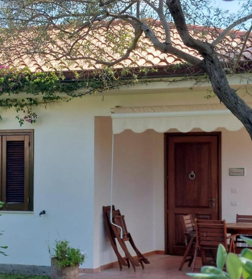 Casa accogliente con veranda, sedie in legno, fiori colorati e alberi ombreggianti.