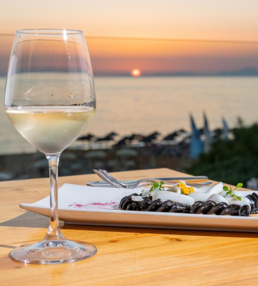 Tramonto sul mare, calice di vino bianco e piatto elegante su tavolo di legno.