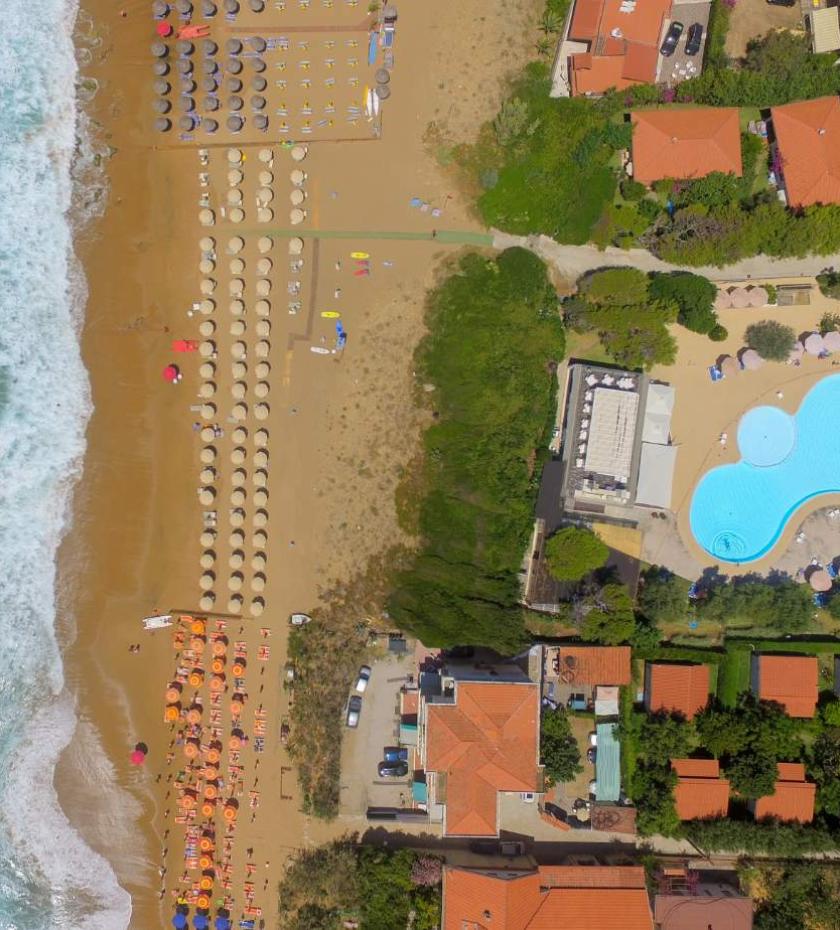 Vista aerea di spiaggia dorata, ombrelloni colorati, piscina azzurra e onde che si infrangono.