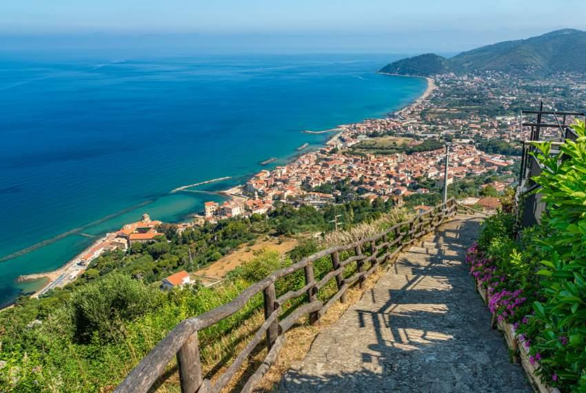 Sentiero panoramico con vista su mare azzurro, colline verdi e un pittoresco borgo costiero.